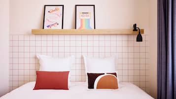 Una cama bien hecha con almohadas blancas y marrones, un estante con obras de arte enmarcadas arriba y una pared con diseño de cuadrícula.