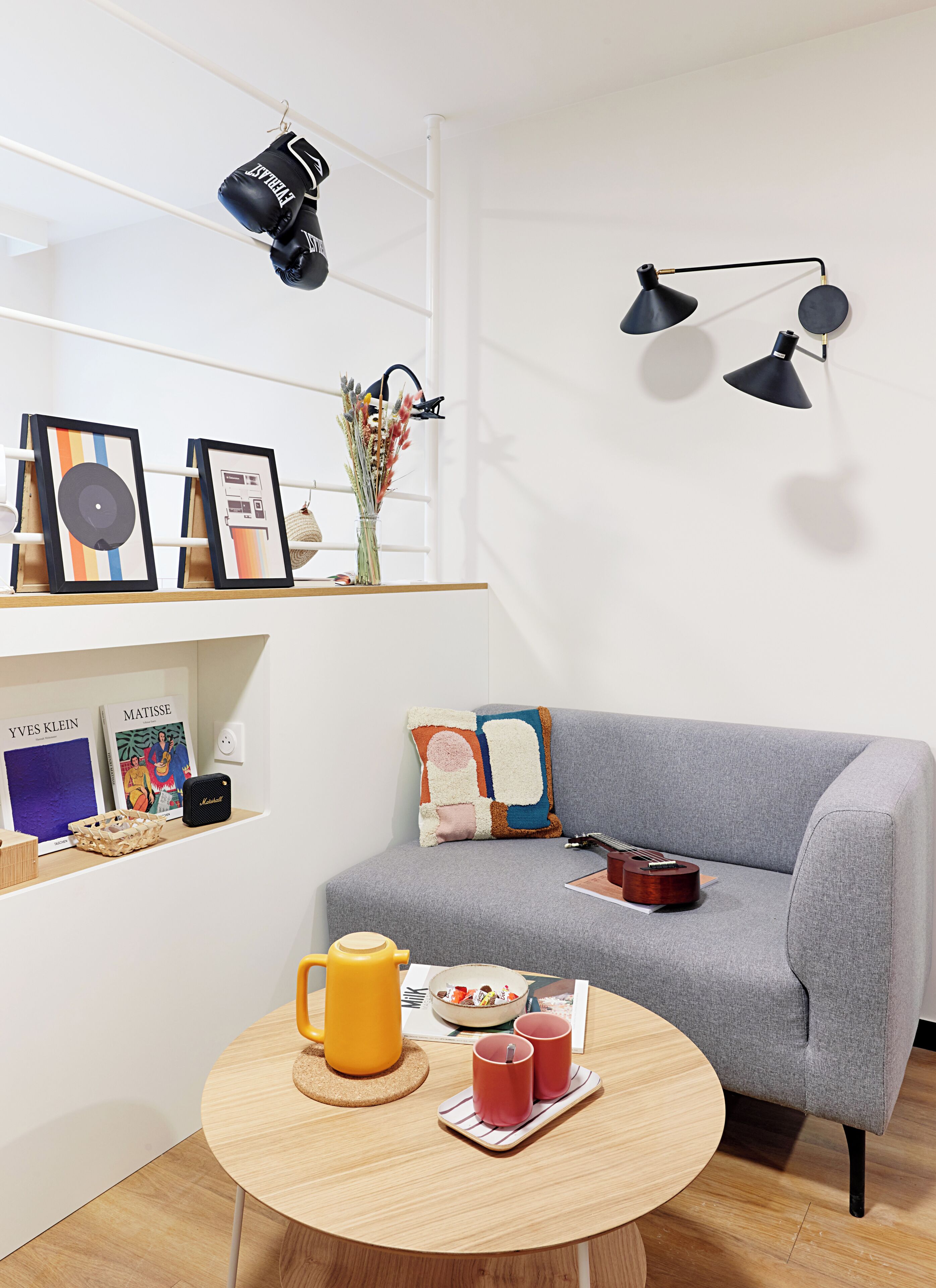 Ein modernes Wohnzimmer mit einem grauen Sofa, einem runden Couchtisch aus Holz und einem Regal mit gerahmten Kunstwerken und einer Vase mit getrockneten Blumen.
