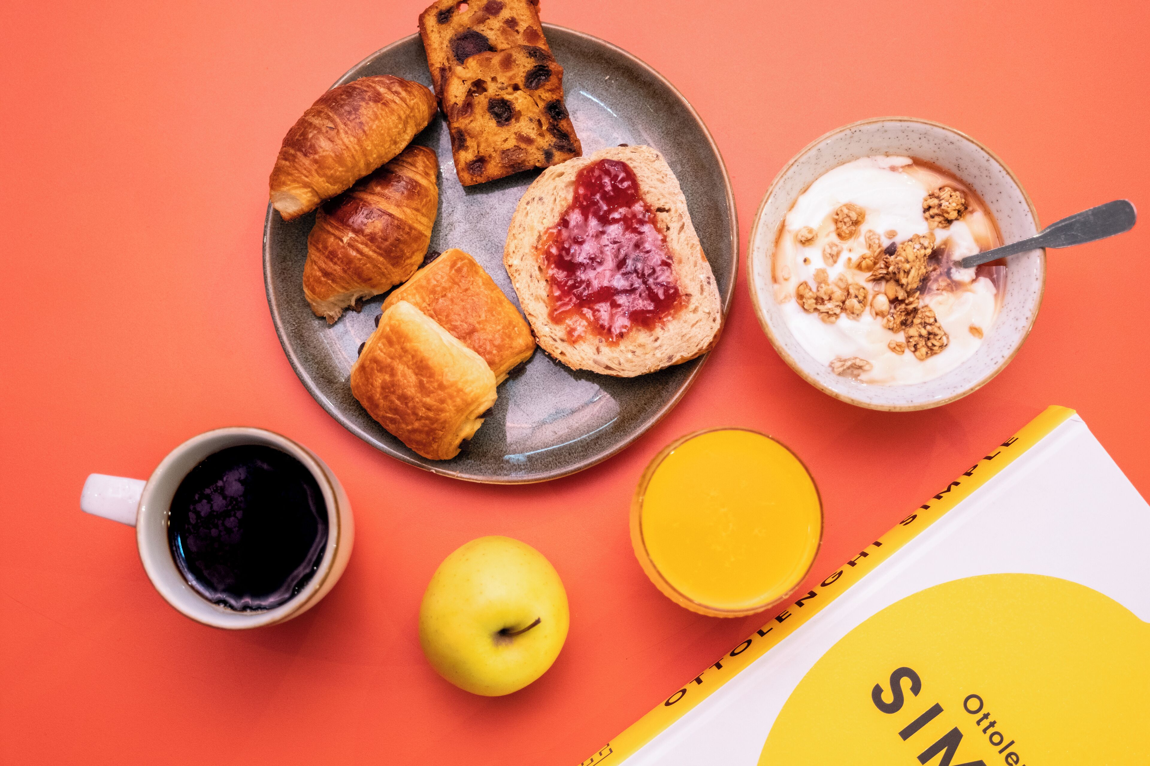 A breakfast setting with croissants, jam, yogurt with granola, a cup of coffee, an apple, and a book on a table.