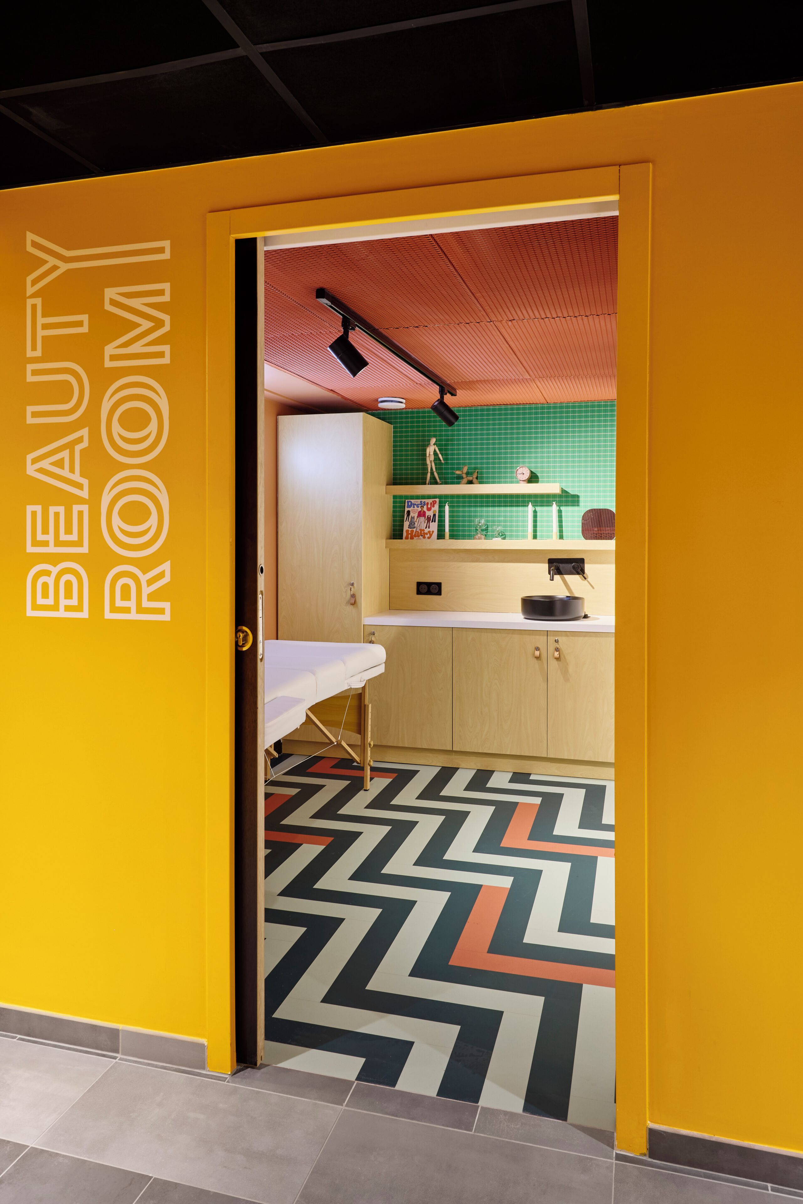 Parede amarela com a inscrição 'BEAUTY ROOM', piso estampado, cozinha compacta com painel verde e teto com vigas aparentes.
