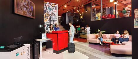 A modern hotel lobby with a red reception desk, a self-check-in station, and a seating area with sofas and a coffee table.