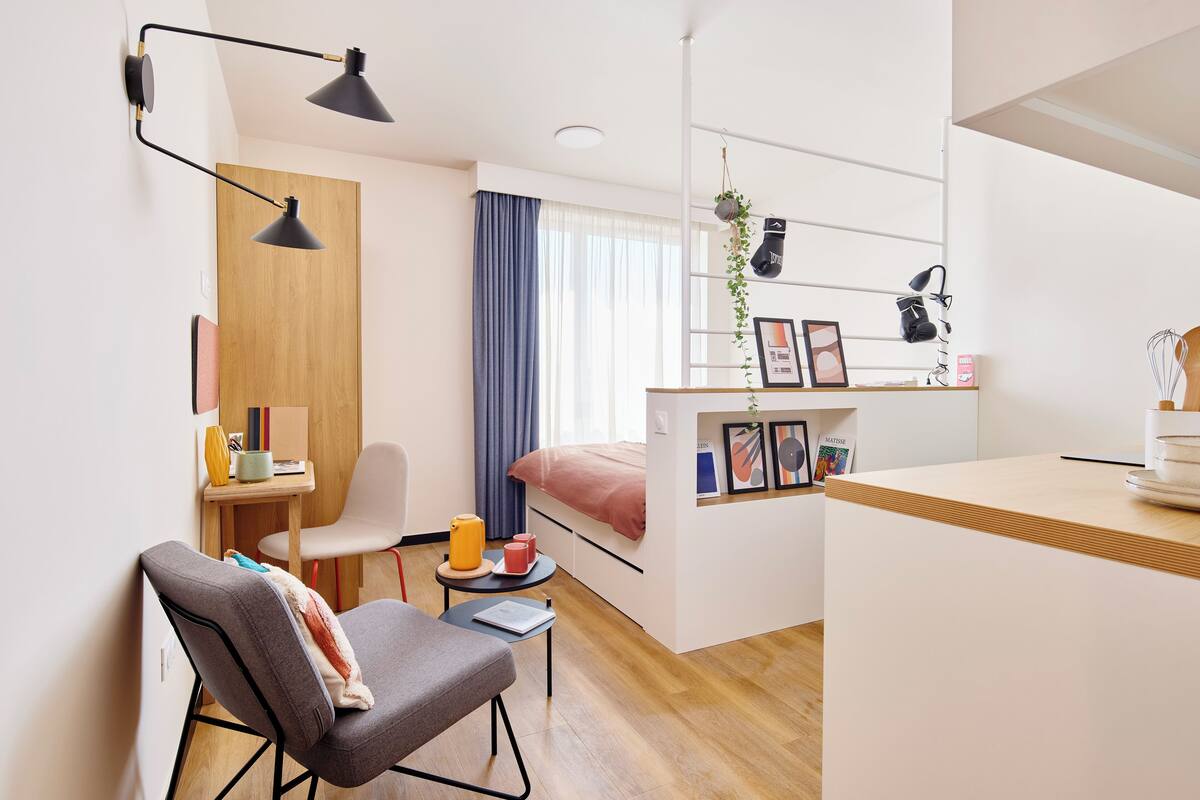 A compact hotel room with a bed, a desk, a chair, a small table, and a shelf with personal items.