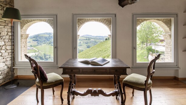 Une pièce avec une table et deux chaises, un livre sur la table et trois fenêtres donnant sur un paysage verdoyant.