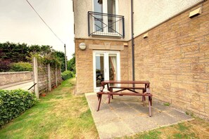 A patio area with a wooden table and benches, a stone wall, and a glass door leading to a balcony. - Ground floor 2-bedroom apartment overlooking the famous James Braid Brora Golf Course. (Highland Council)