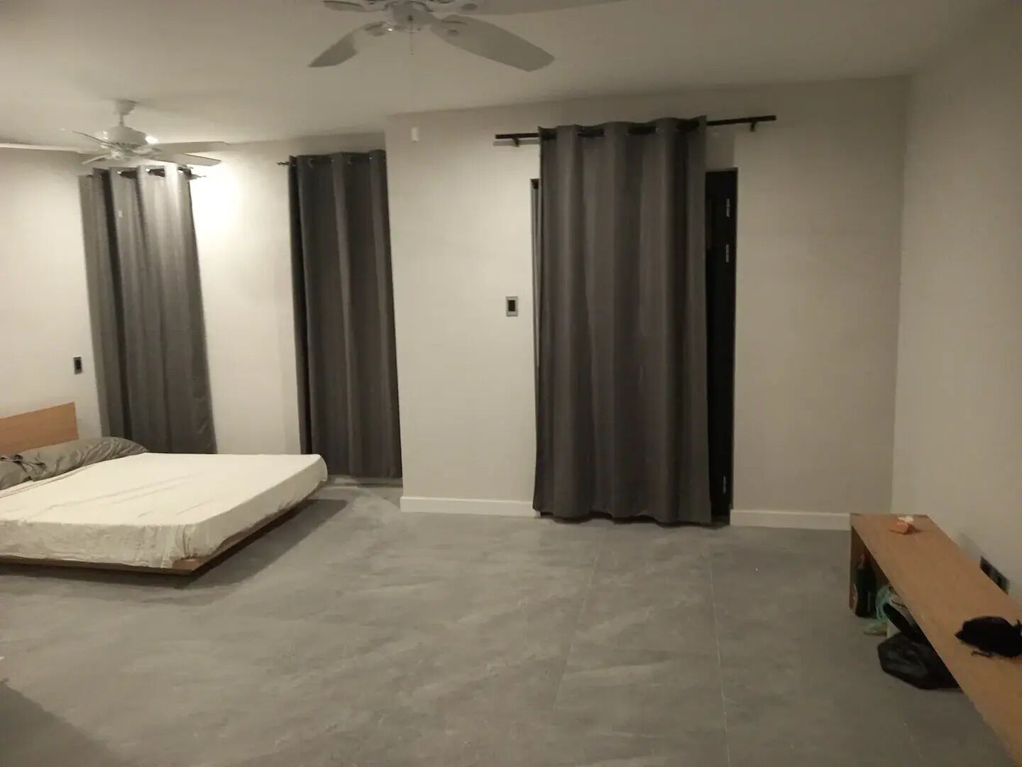 Un dormitorio con cama, ventilador de techo, cortinas grises y un banco de madera.
