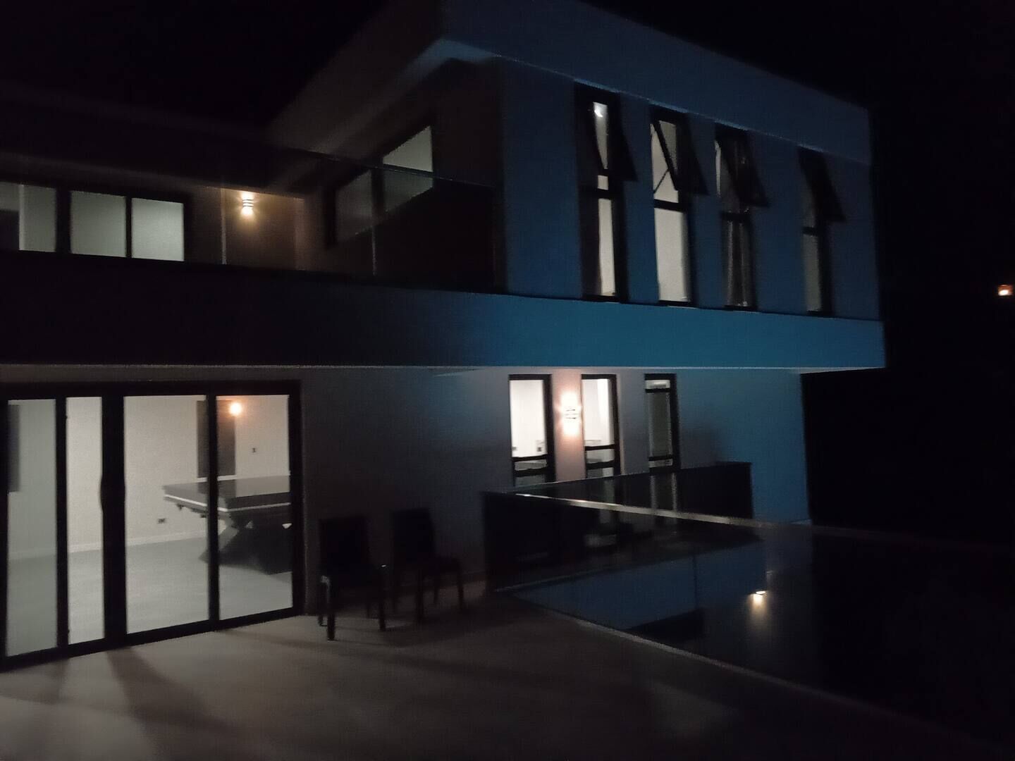 En moderne bygning med to etager, en glasfacade, en balkon og et spiseområde med stole og et bord.