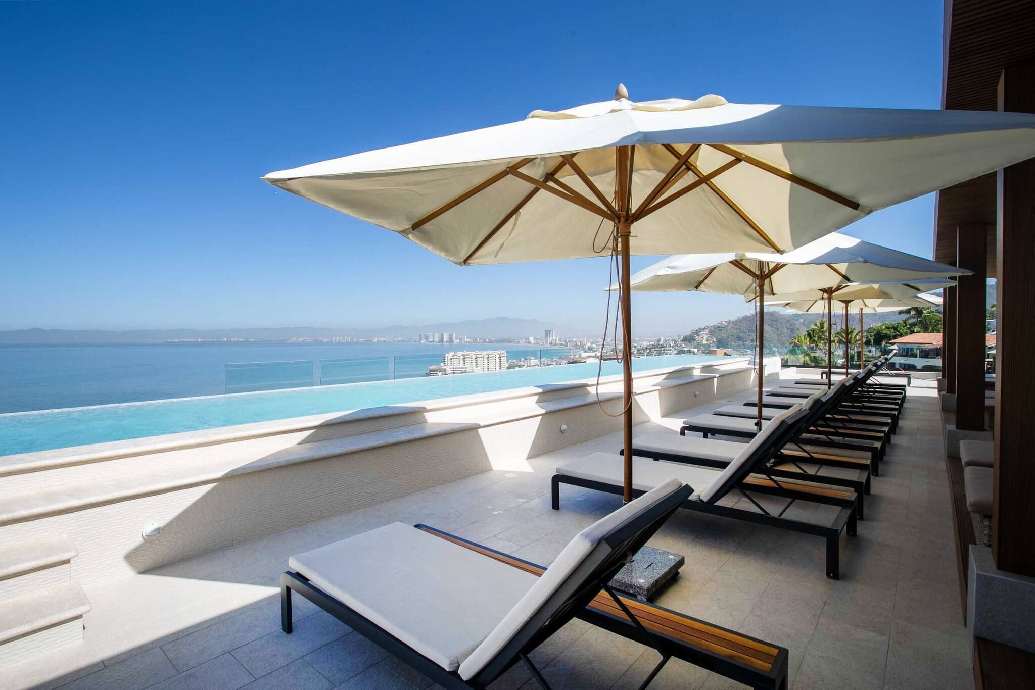 Zona de piscina con sillones de descanso y sombrillas, con vistas a un mar azul y una ciudad a lo lejos.