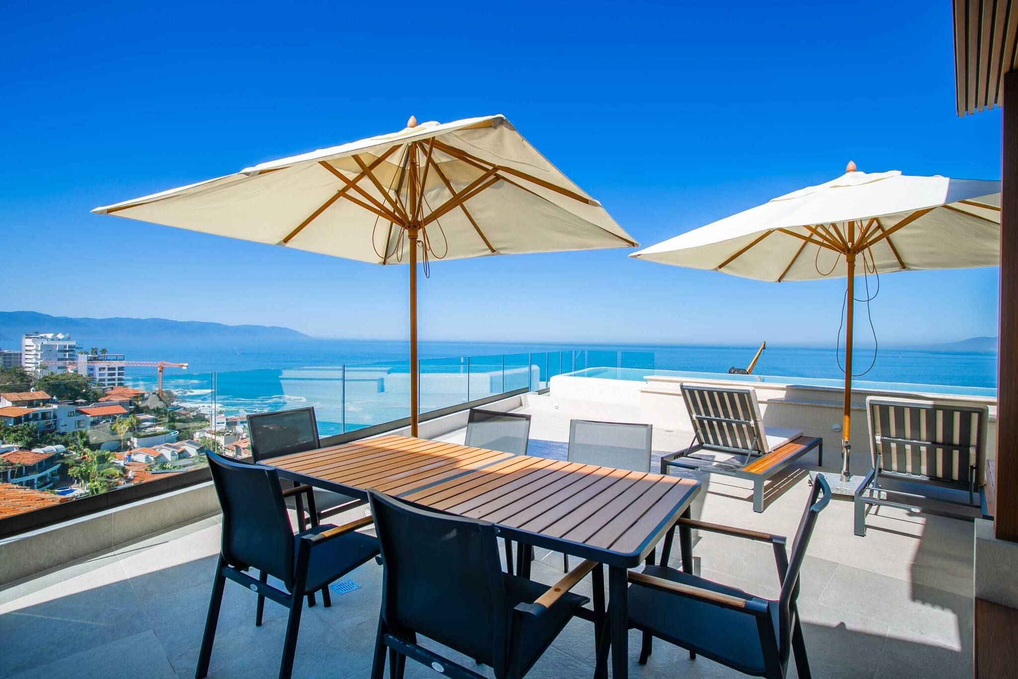 A terrace with a wooden table and chairs, two large umbrellas, and a view of the sea.