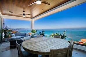A spacious balcony with a wooden table and chairs, overlooking a clear blue sea and distant cityscape.