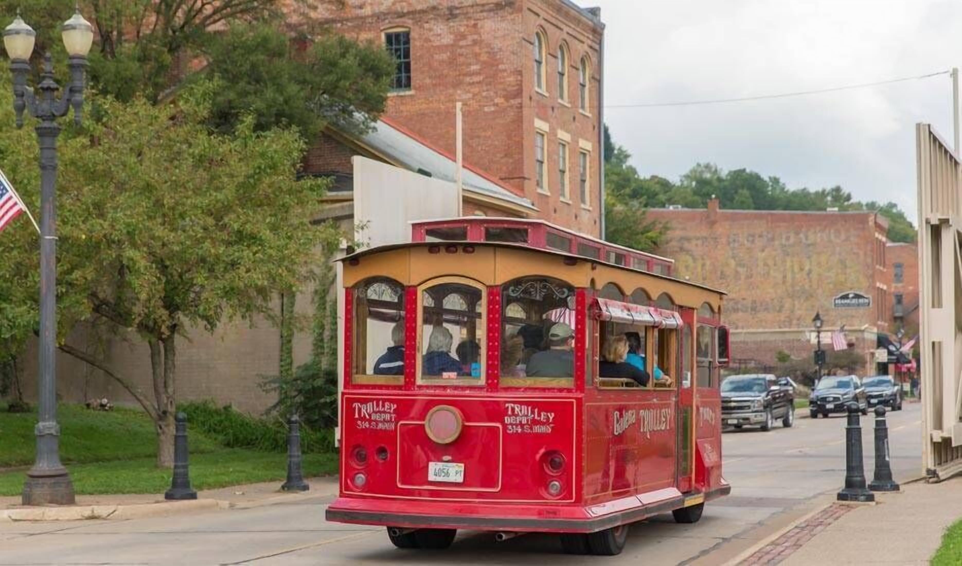 A red trolley with "Trolley Depot" written on the side, traveling down a street with brick buildings and parked cars.