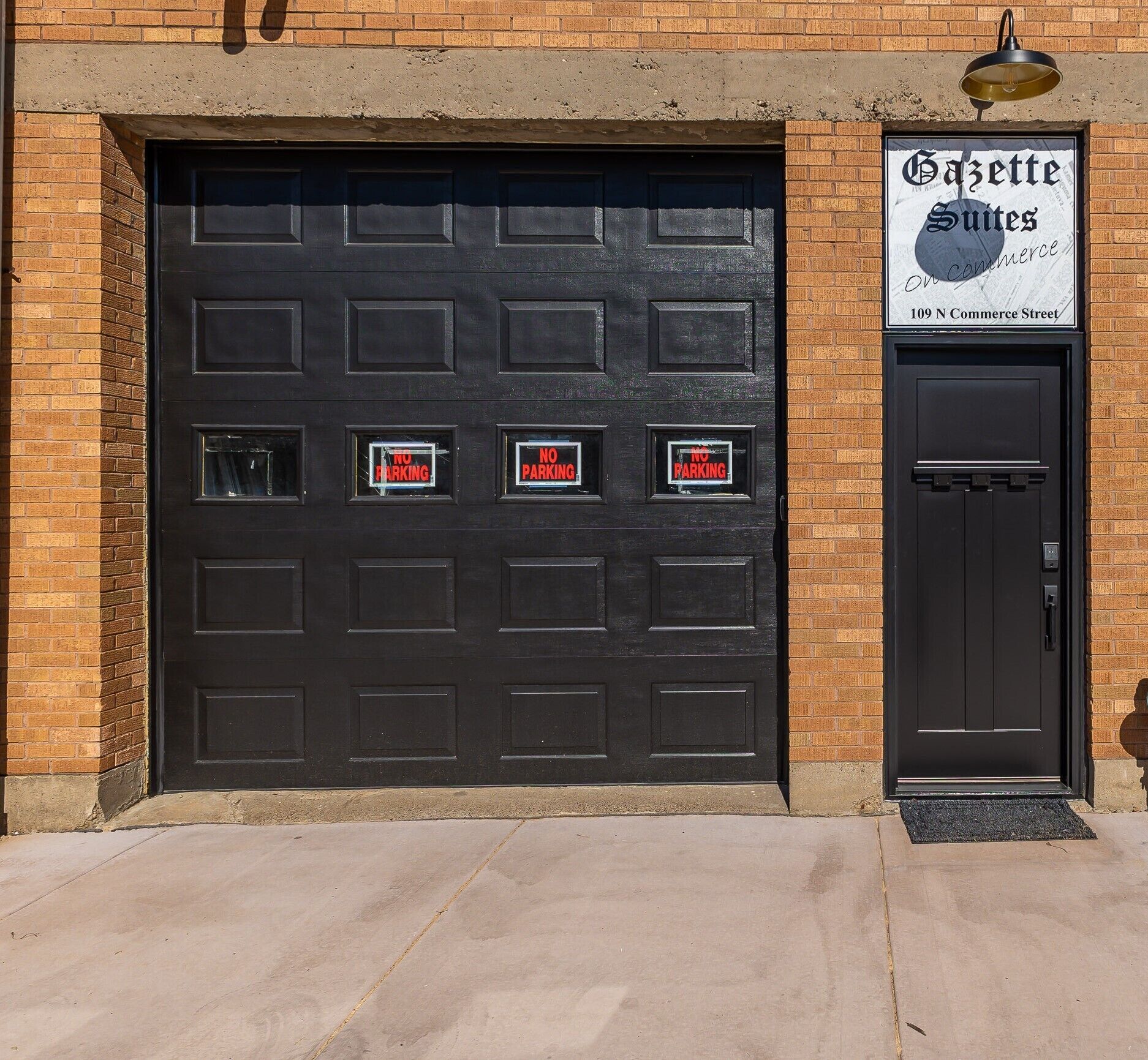 Un portone da garage nero con diverse finestre, ognuna con un cartello che indica 'NO PARKING'. Alla destra, una porta nera con un cartello sopra che recita 'Gazette Suites 109 N Commerce Street'.