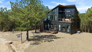 A modern two-story cabin with large windows, a deck, and a gravel driveway surrounded by pine trees. - 54BD/3BTH Serenity Skyline Sleeps 14, Hot Tub and more! (Broken Bow)