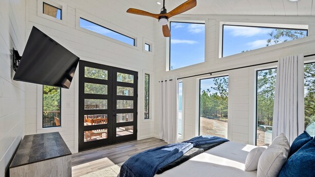 A bedroom with a bed, a TV mounted on the wall, and large windows. - 54BD/3BTH Serenity Skyline Sleeps 14, Hot Tub and more! (Broken Bow)
