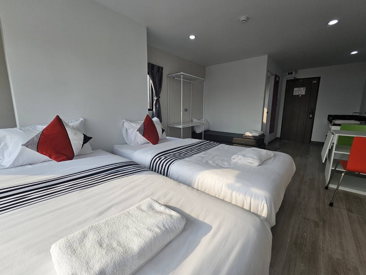 Ein Hotelzimmer mit zwei Betten, jeweils mit weißer Bettwäsche und roten Kissen. Ein kleiner Tisch mit einem Stuhl sowie ein Schreibtisch mit Stuhl sind ebenfalls vorhanden.
