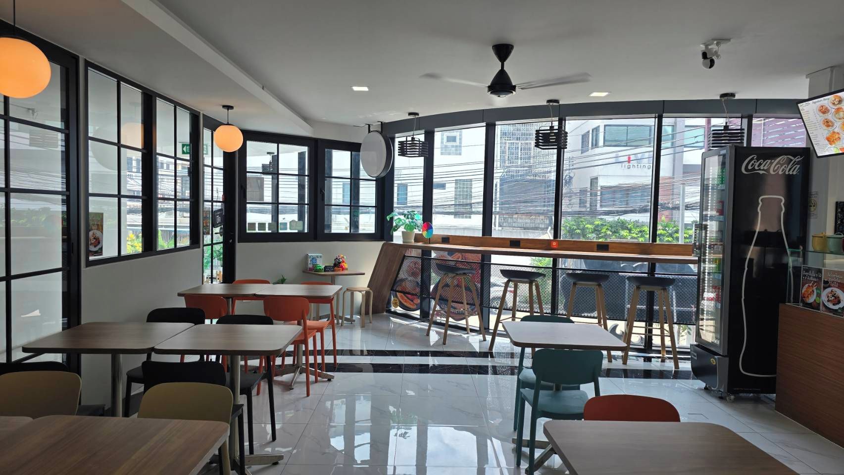 A modern cafe interior with tables and chairs, a large window offering a city view, and a Coca-Cola refrigerator.