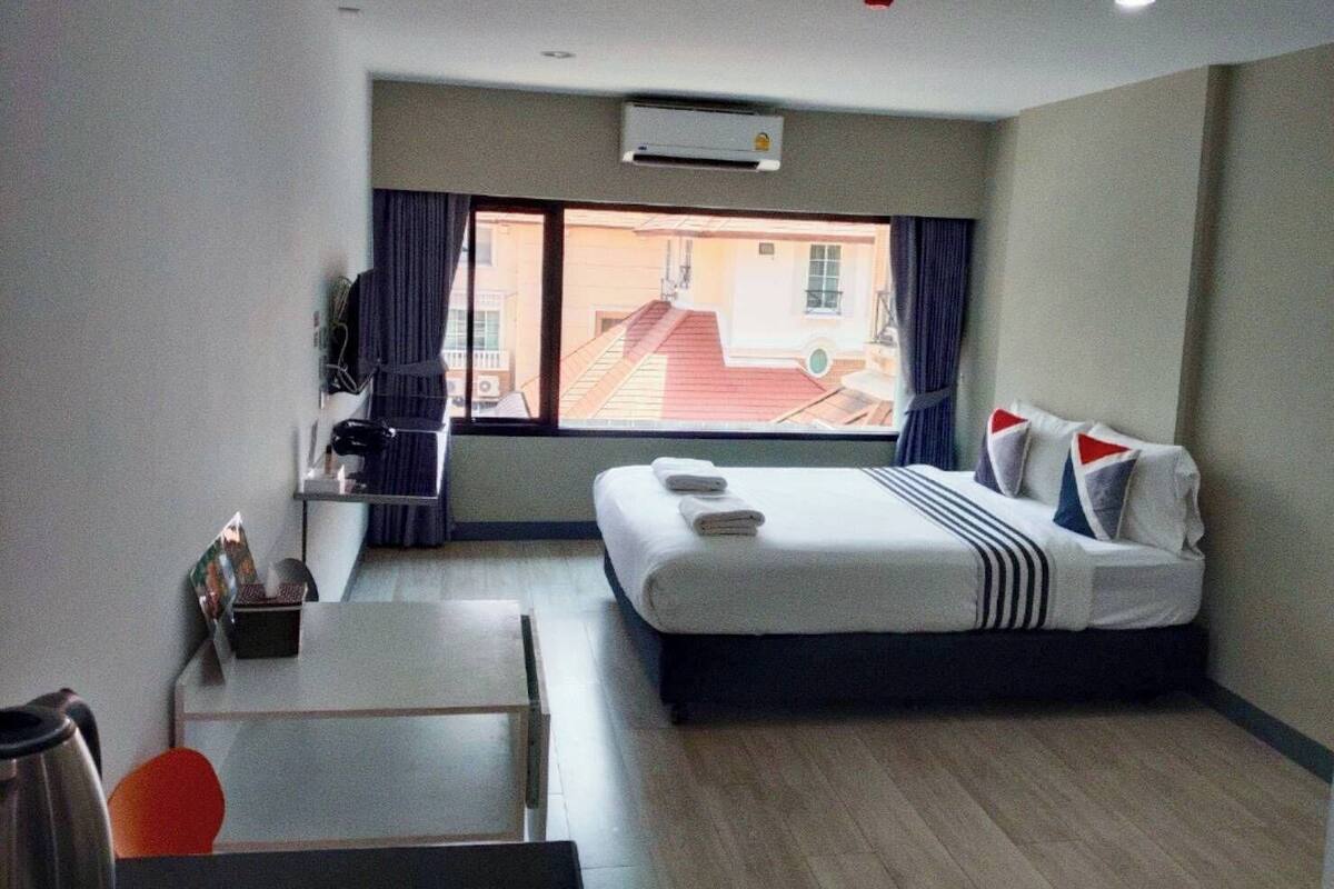 A hotel room with a bed, a desk, a chair, a window with curtains, and an air conditioning unit.