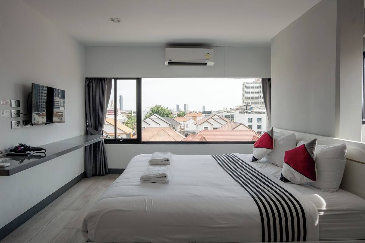 A modern hotel room with a large bed, a flat-screen TV mounted on the wall, and a city view through the window.