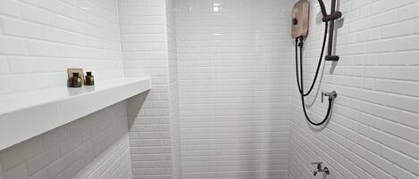 A small, white-tiled bathroom with a shower, a shelf with toiletries, and a dark floor.