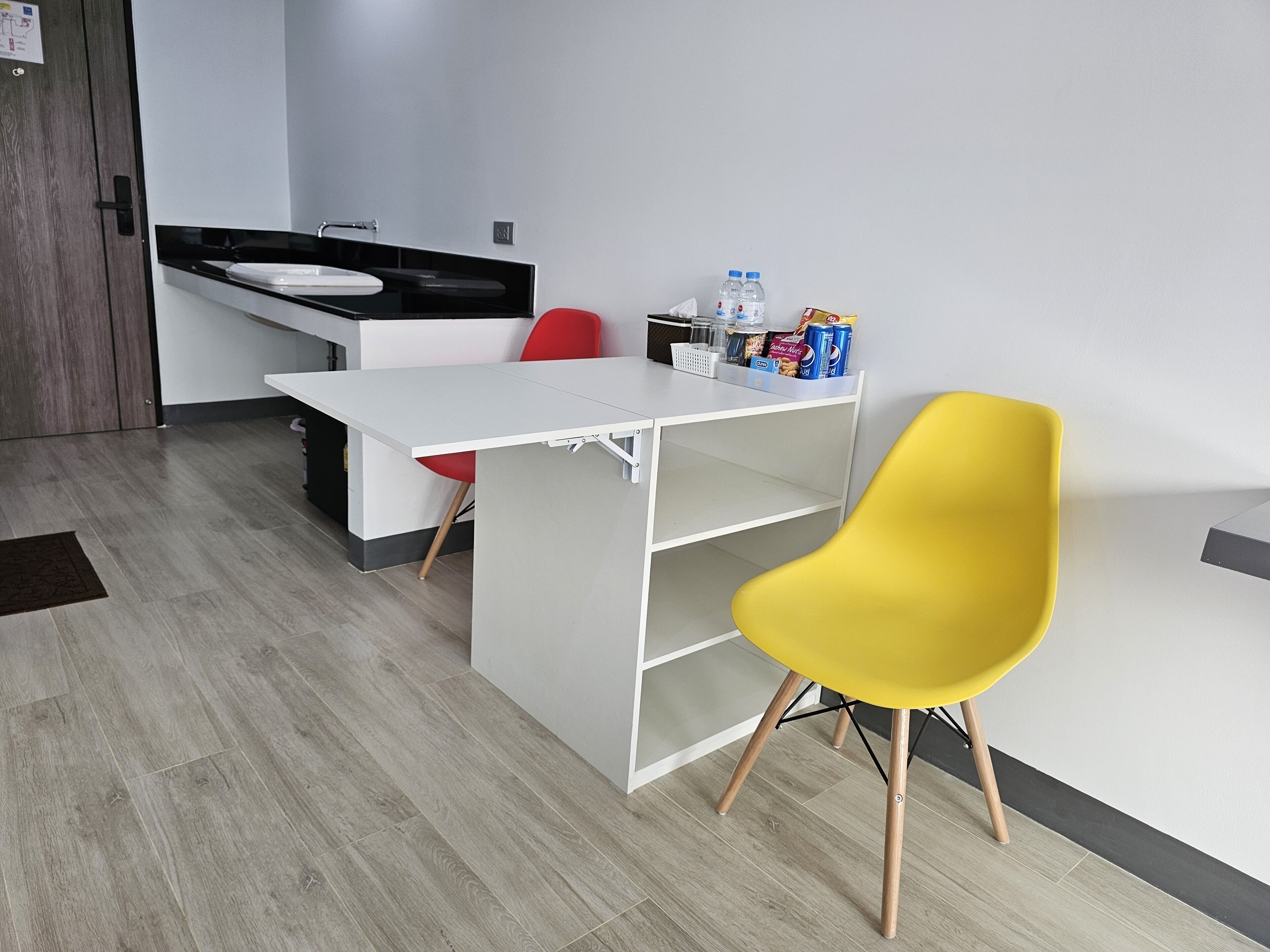 A small kitchenette with a white countertop, a sink, and a red chair. There is a yellow chair in front of a white shelf with snacks and drinks.