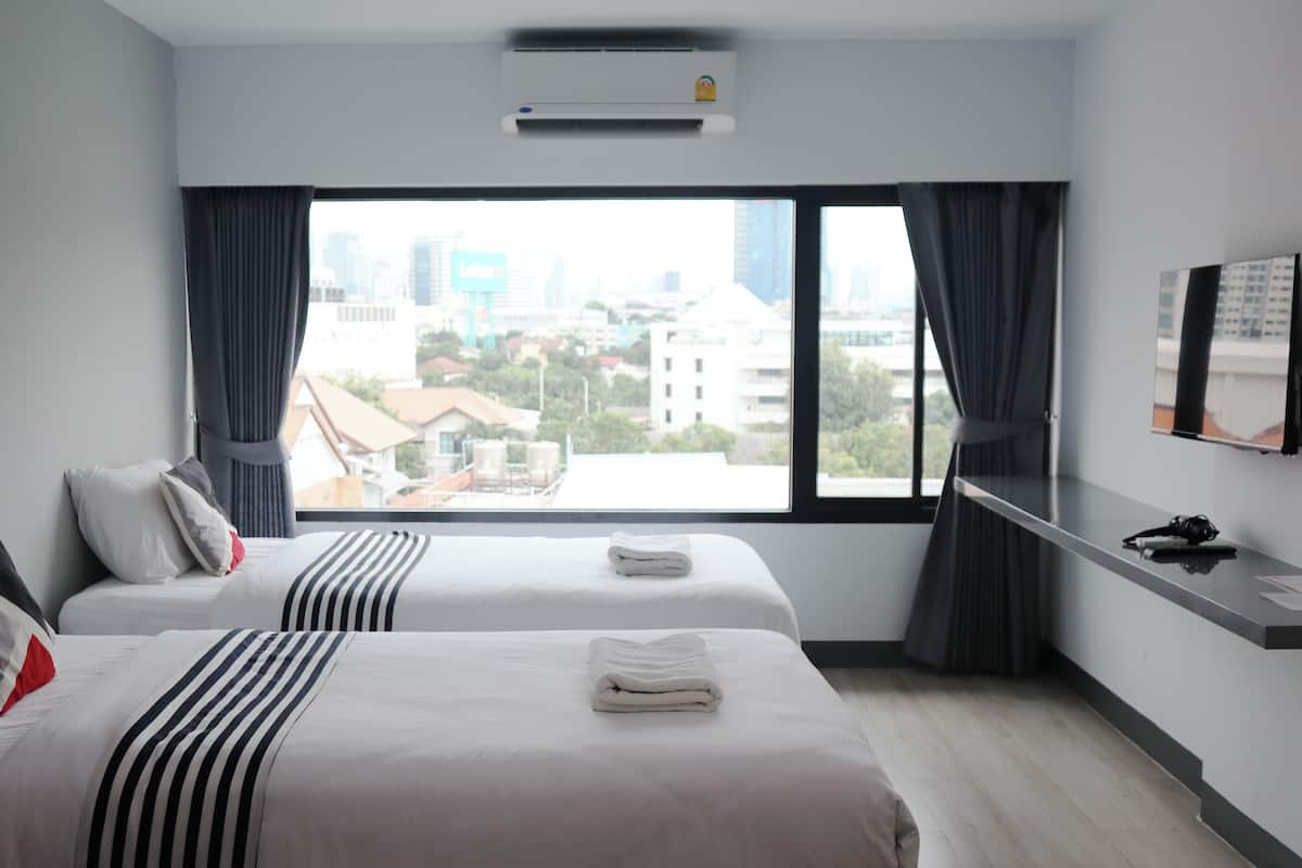 A hotel room with two beds, a large window with a city view, a flat-screen TV on the wall, and a small desk with a chair.