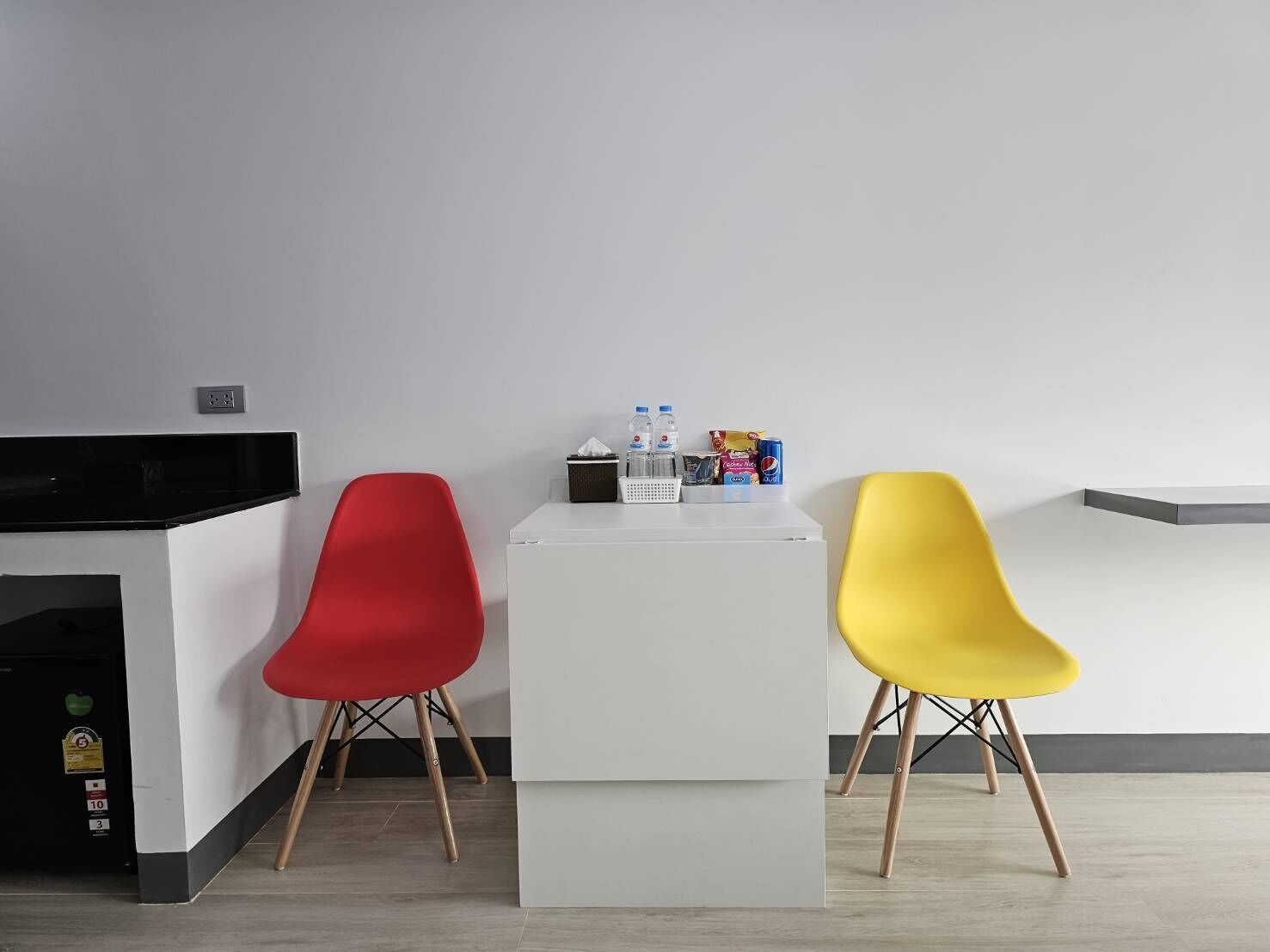 A small kitchenette with a microwave, a white table with a tissue box, water bottles, and snacks. Two chairs, one red and one yellow, are placed in front of the table.