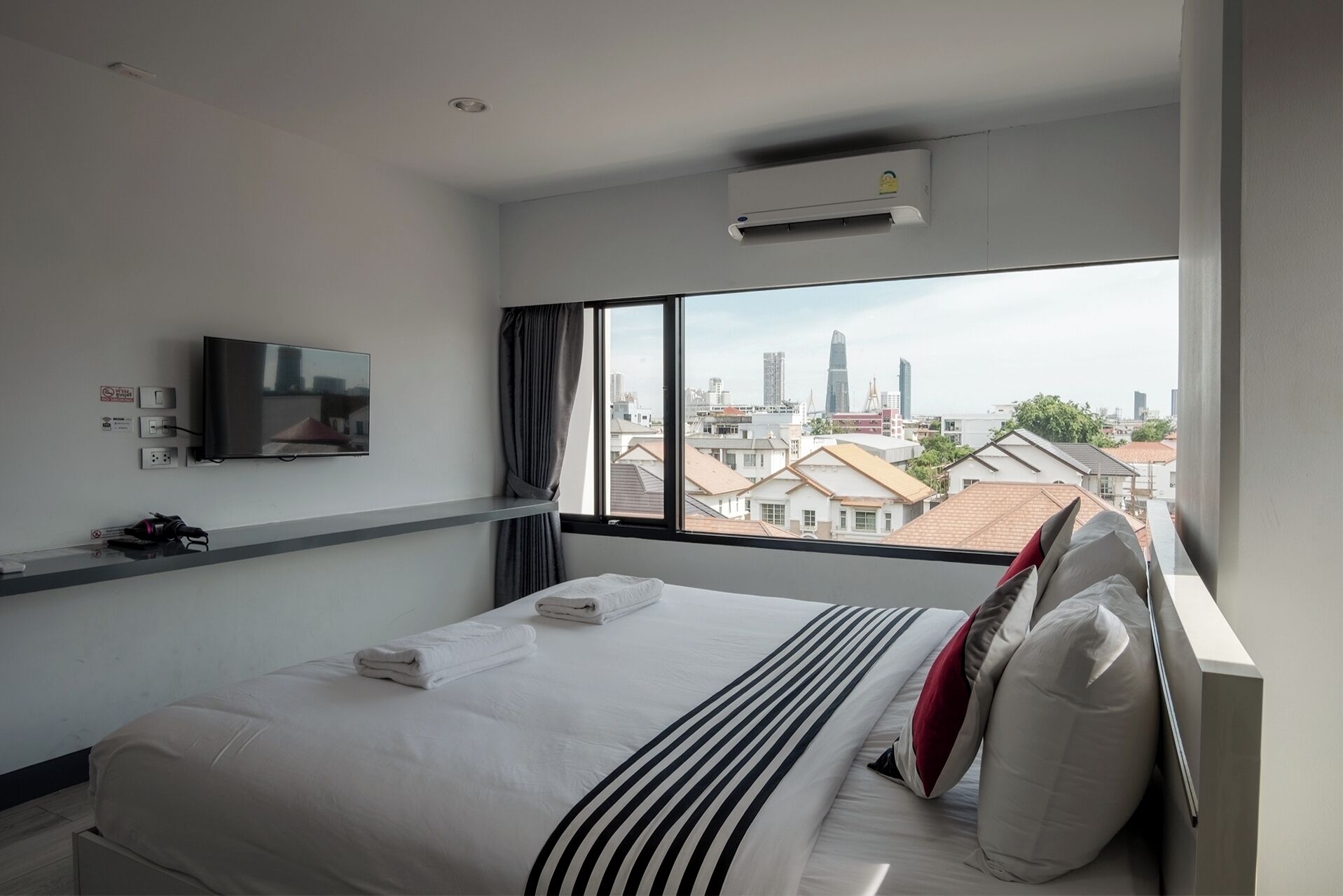 A hotel room with a bed, a flat-screen TV mounted on the wall, and a large window offering a city view.