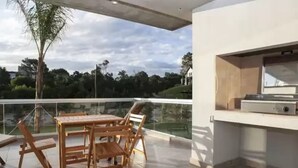 An outdoor patio with a wooden table and chairs, a grill, and a glass railing overlooking a green landscape. - Nova Pampa apart&spa (Mar Azul)