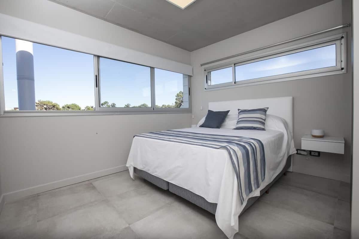 A bedroom with a bed, two windows, a nightstand, and a view of the outside.