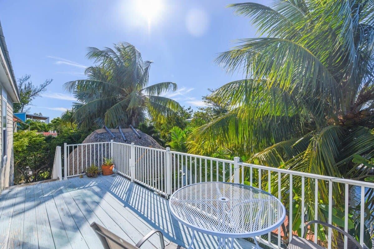 A wooden deck with a metal table and white railing, surrounded by palm trees and greenery.