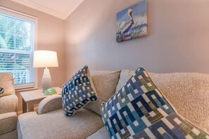 A cozy living room with a beige sofa, patterned cushions, a painting of a pelican, and a lamp on a side table.