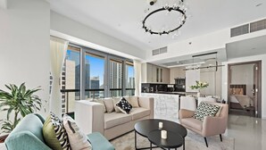 A modern living room with a sofa, armchair, and coffee table, large windows, and a kitchen in the background.