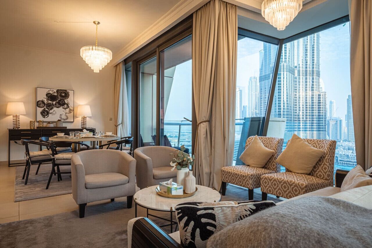 A modern hotel room with a large window offering a city skyline view, a dining area with a round table and chairs, and a cozy seating area with patterned cushions.