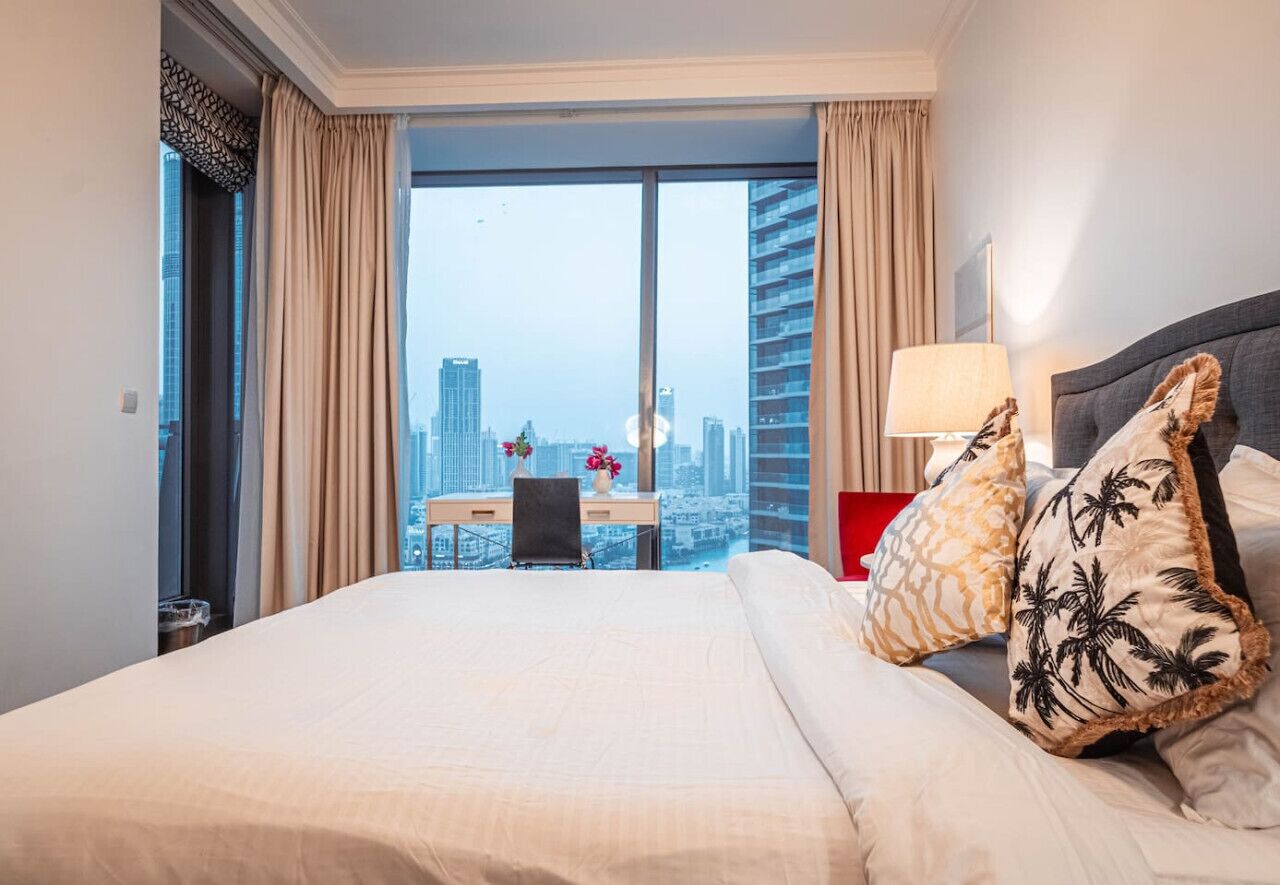A hotel room with a large bed, a desk, and a view of a cityscape through floor-to-ceiling windows.