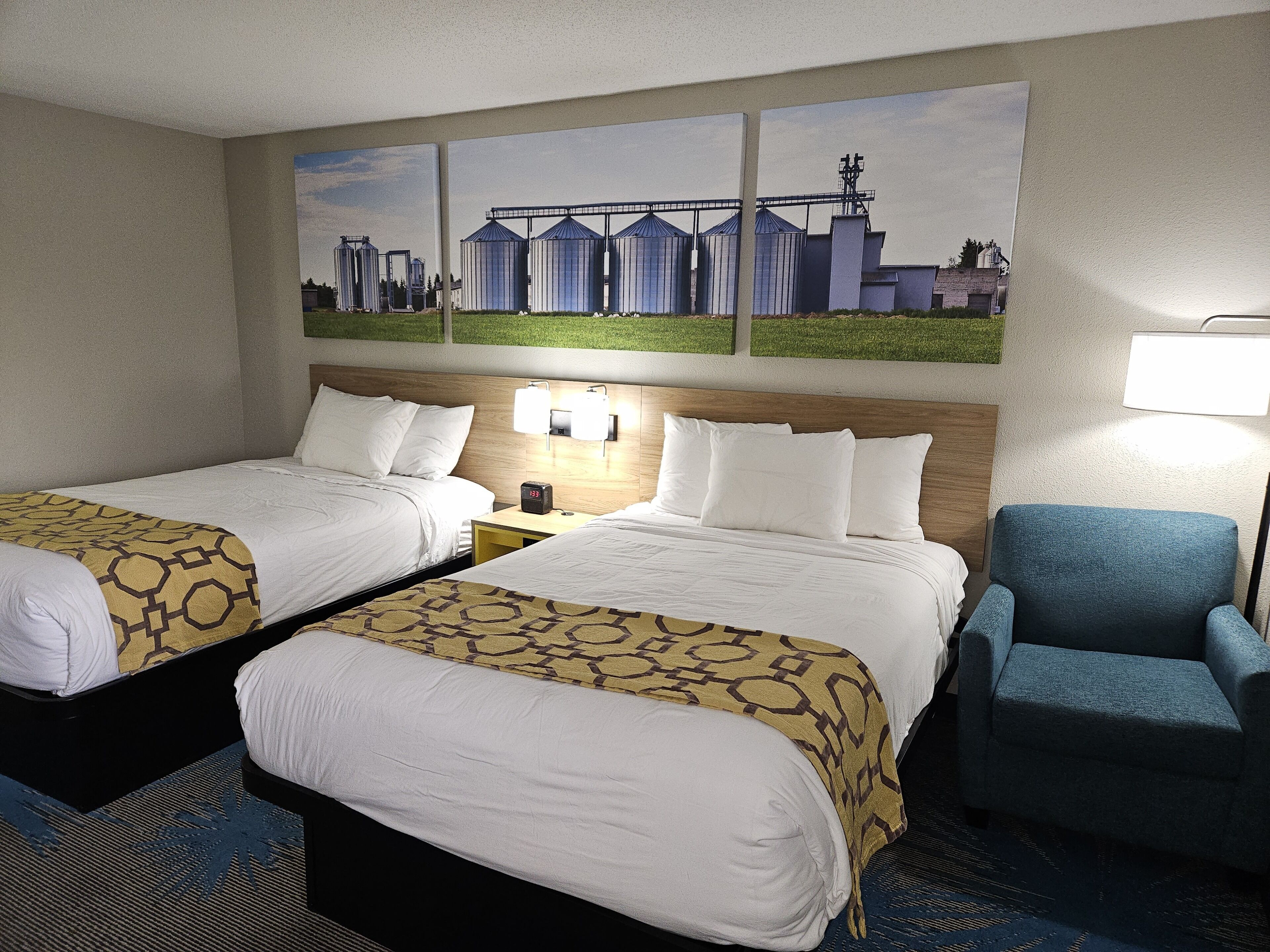 A hotel room with two beds, a nightstand with a clock, a chair, and a painting of industrial buildings on the wall.