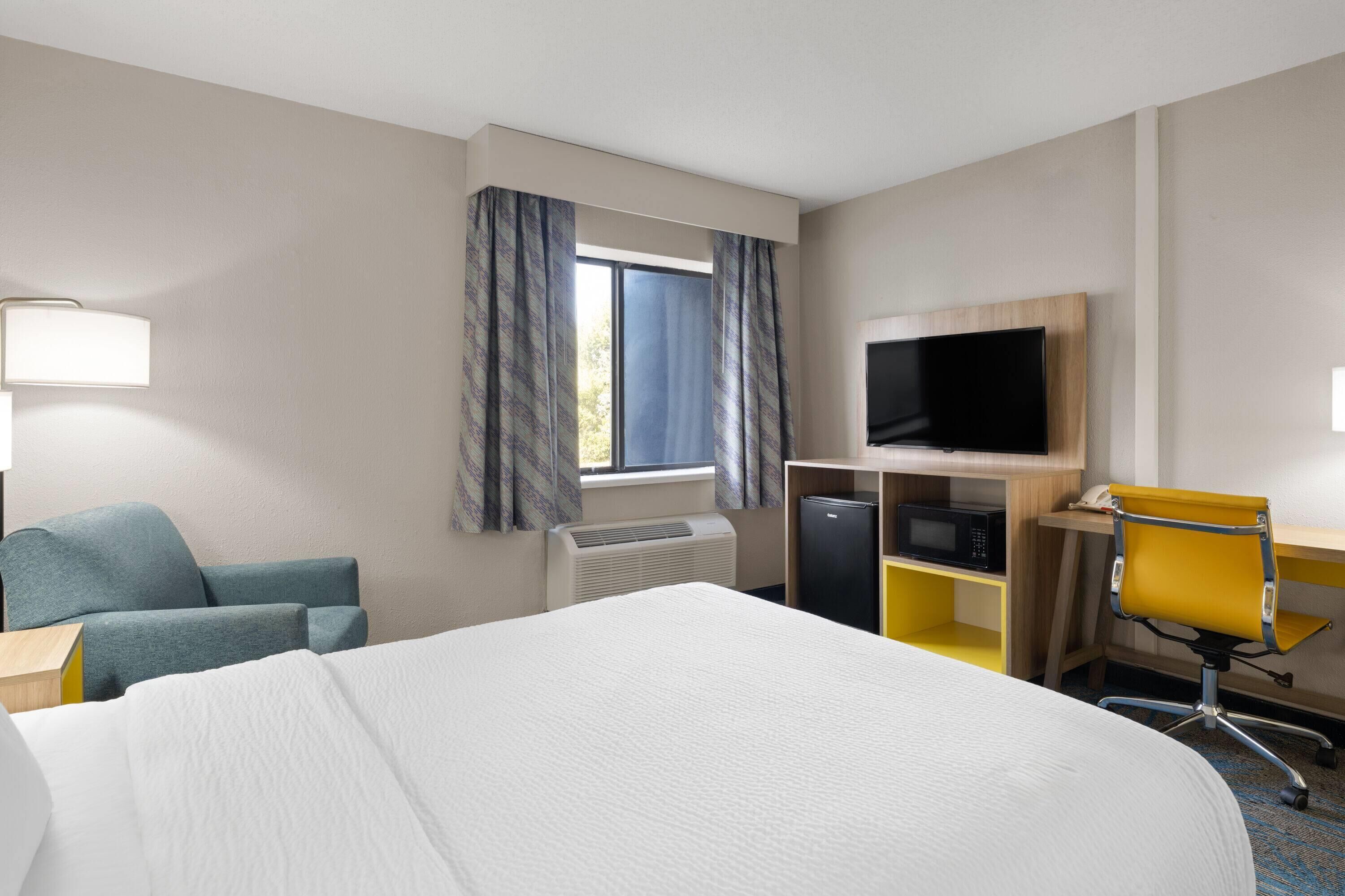 Habitación de hotel con cama, escritorio con computadora, televisor, silla y ventana con cortinas.