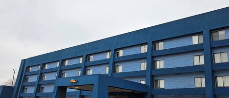 Un edificio grande y azul con varios pisos, una entrada cubierta y un estacionamiento con varios coches.