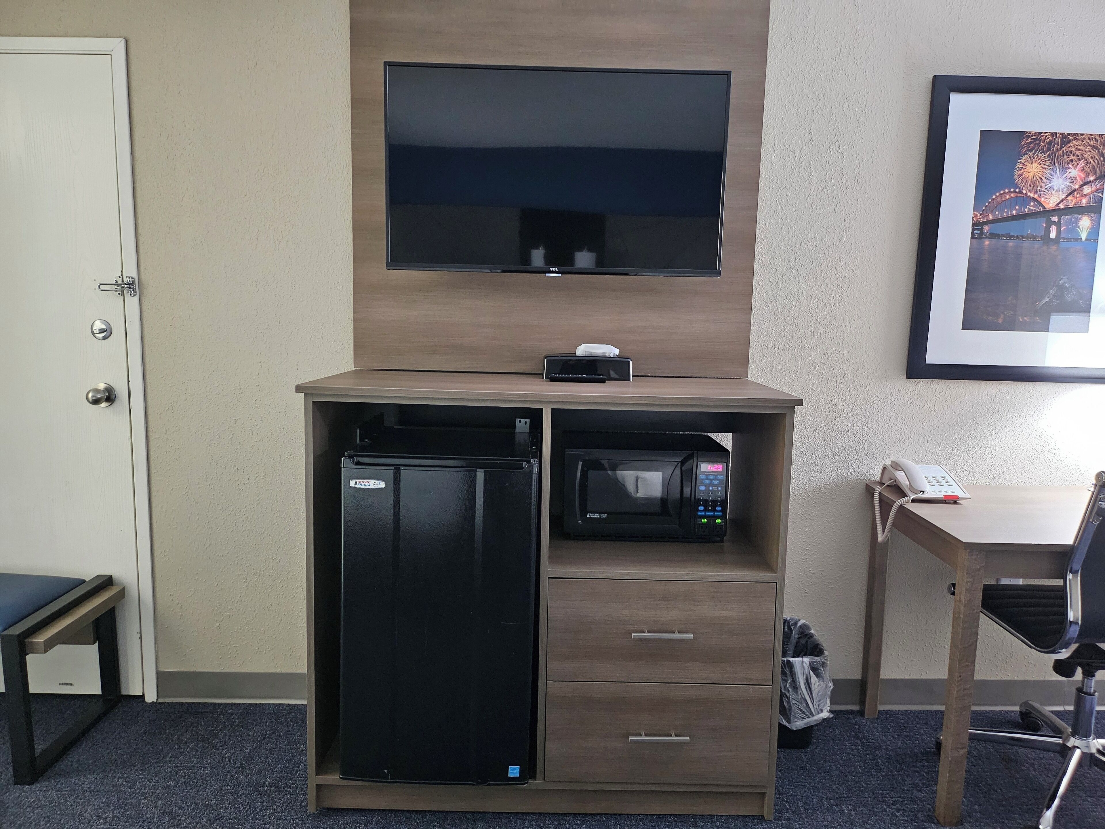 A compact hotel room with a built-in TV, a microwave, a mini fridge, a desk with a phone, and a framed picture on the wall.