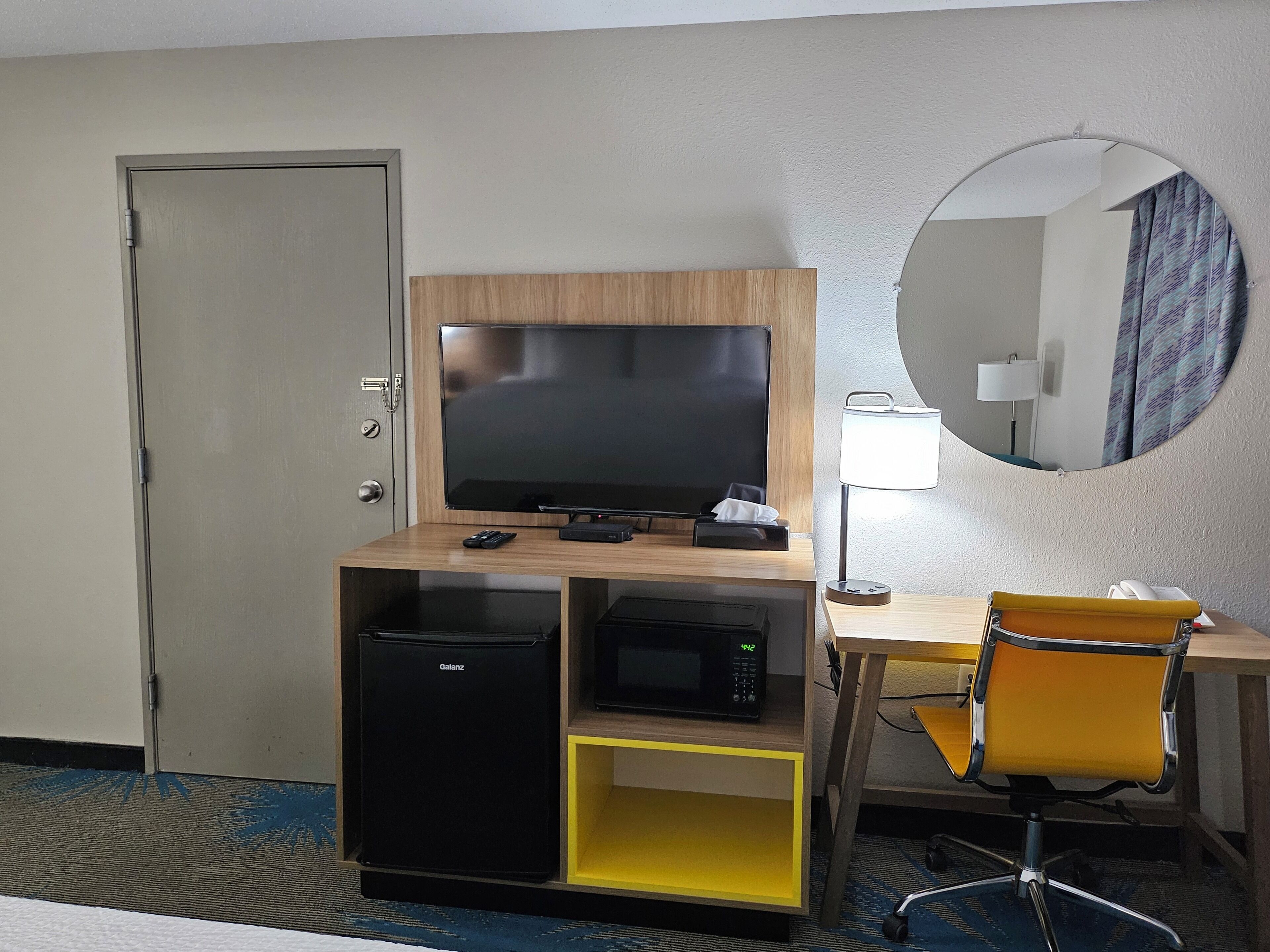 Et hotelværelse med fjernsyn, mikrobølgeovn, lille køleskab, skrivebord med lampe og stol.
