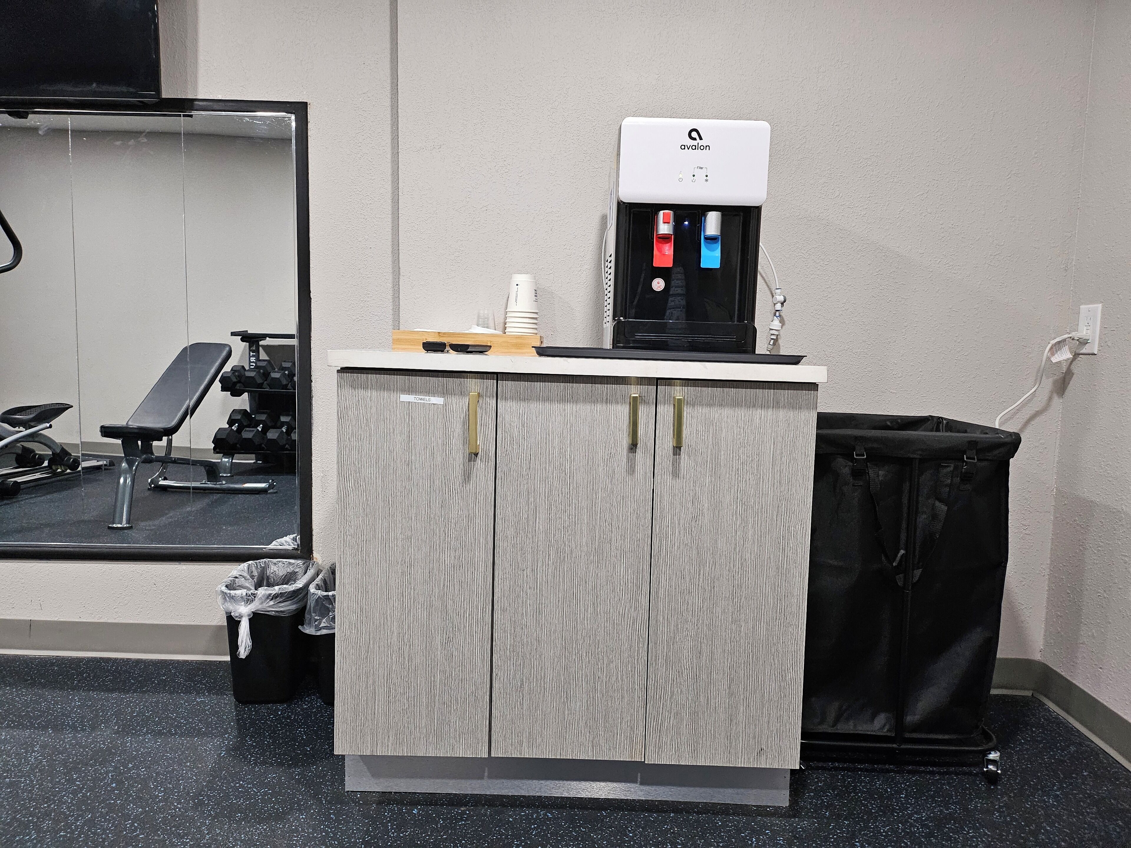 Ein Fitnessraum mit Wasserspender, Spiegel, Bank und Mülleimer.