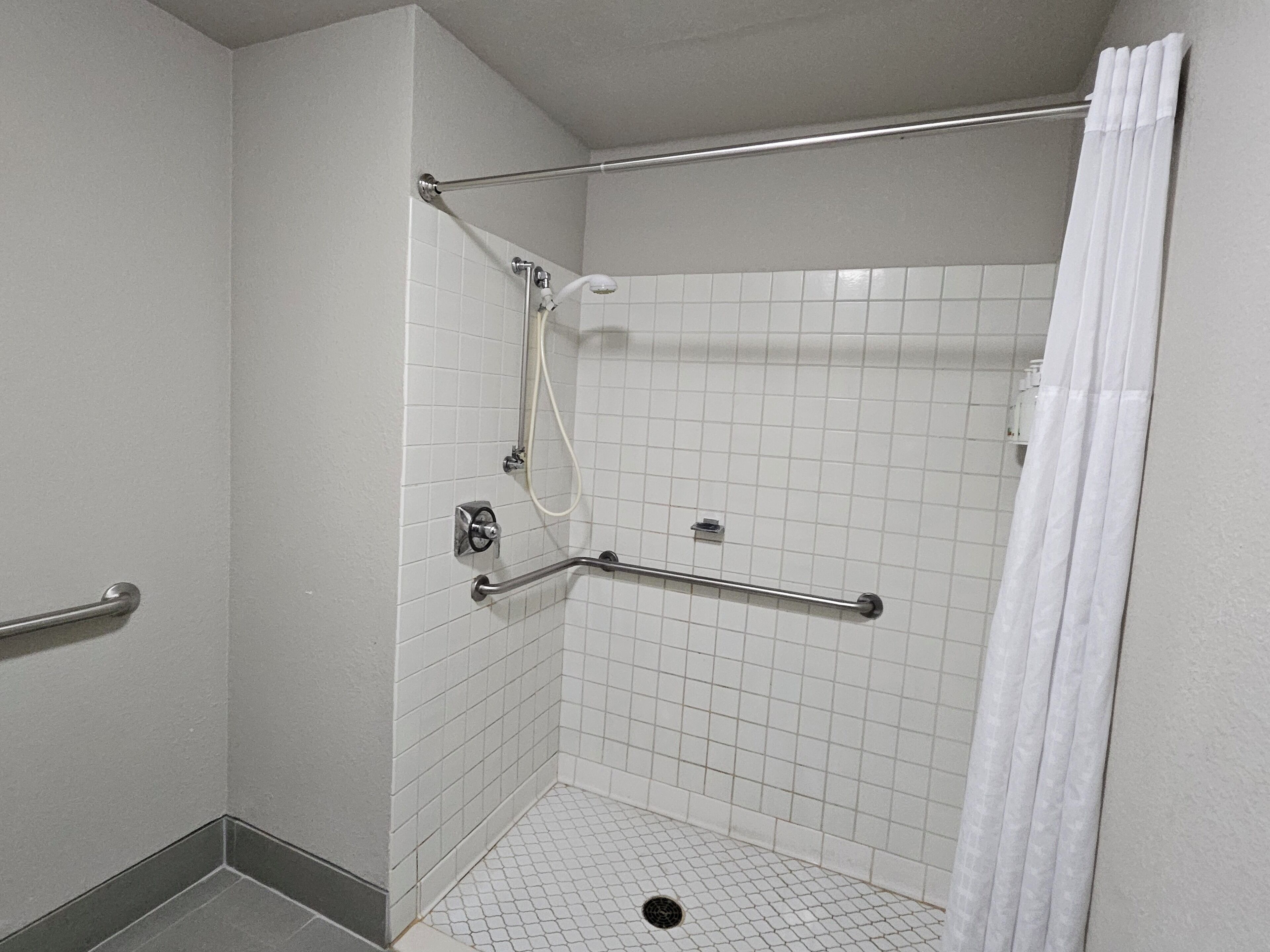 A small, tiled shower area with a white curtain, grab bars, and a handheld showerhead.