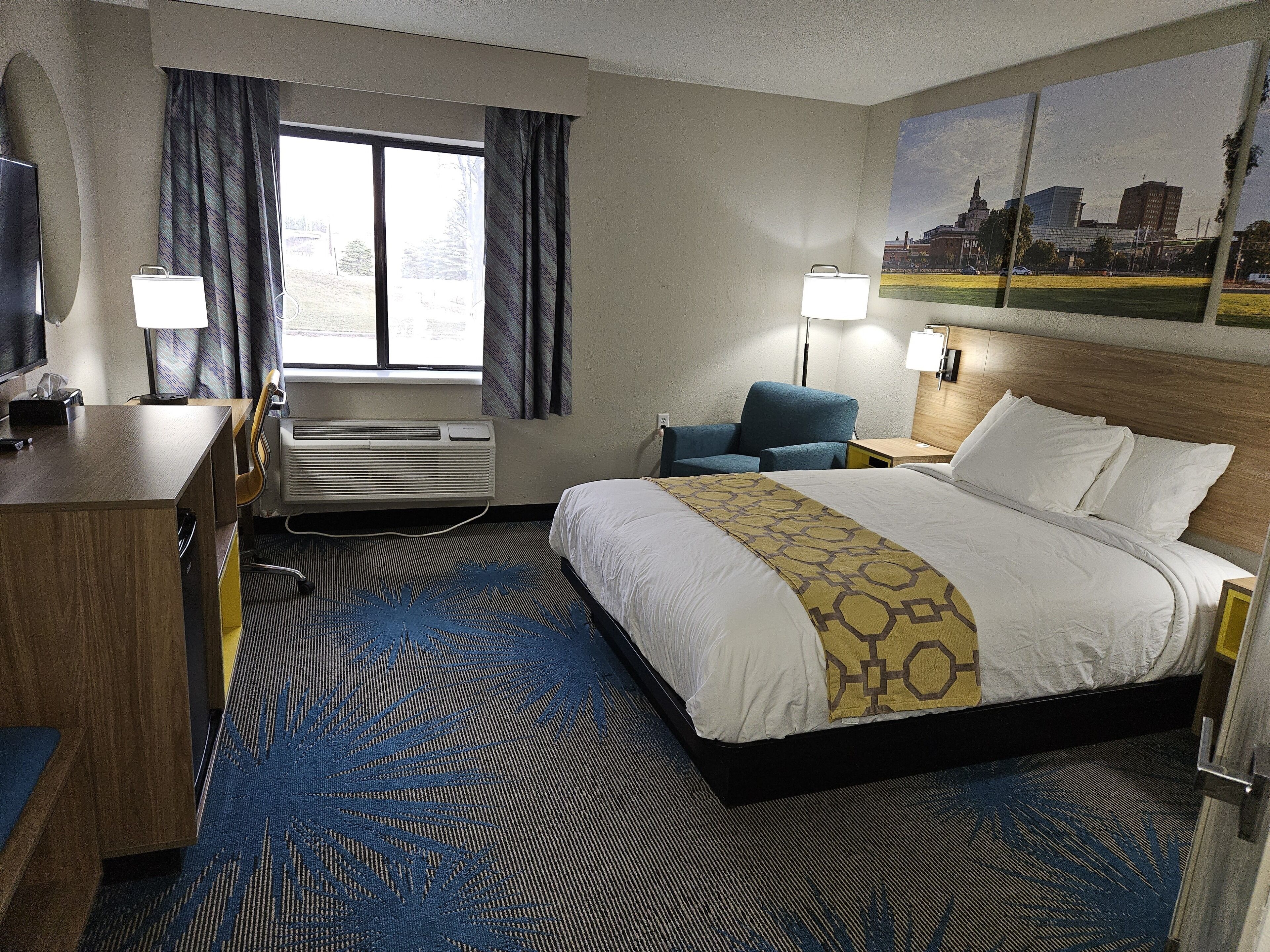 A hotel room with a bed, a desk, a chair, a lamp, and a window with curtains.