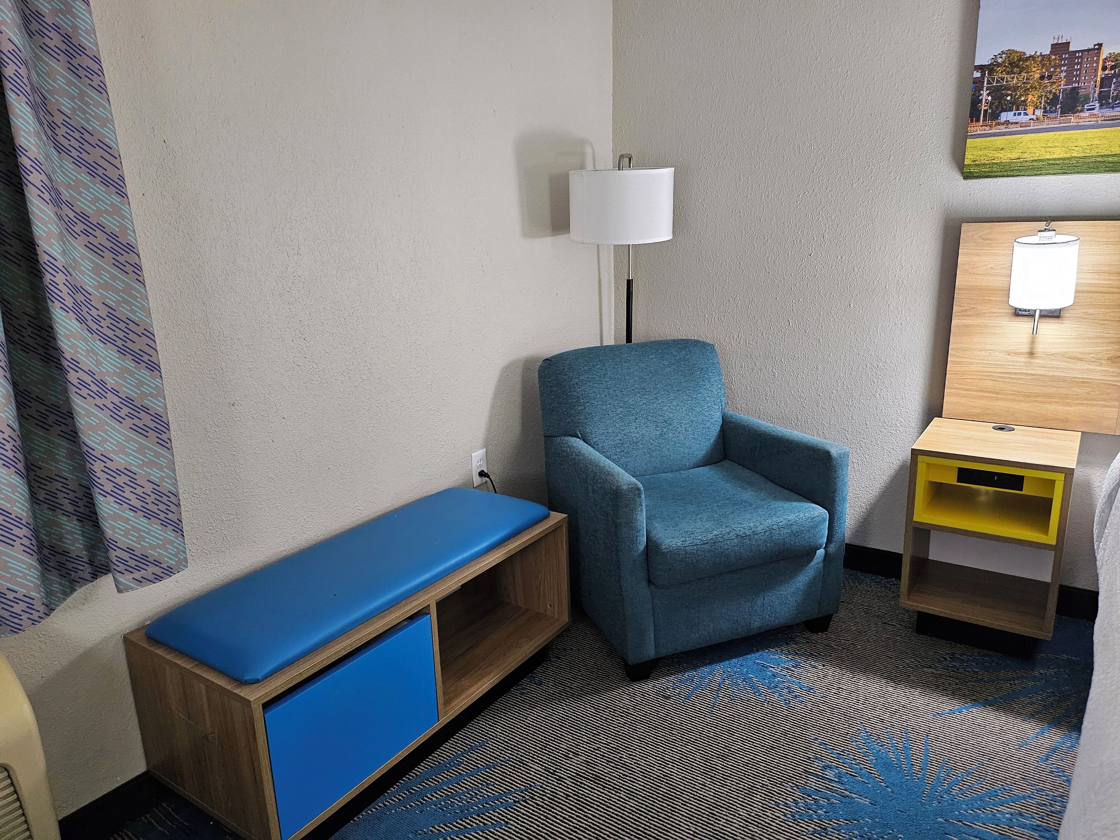 Una habitación de hotel con un sillón azul, una mesita de noche de madera con una lámpara y un banco azul.