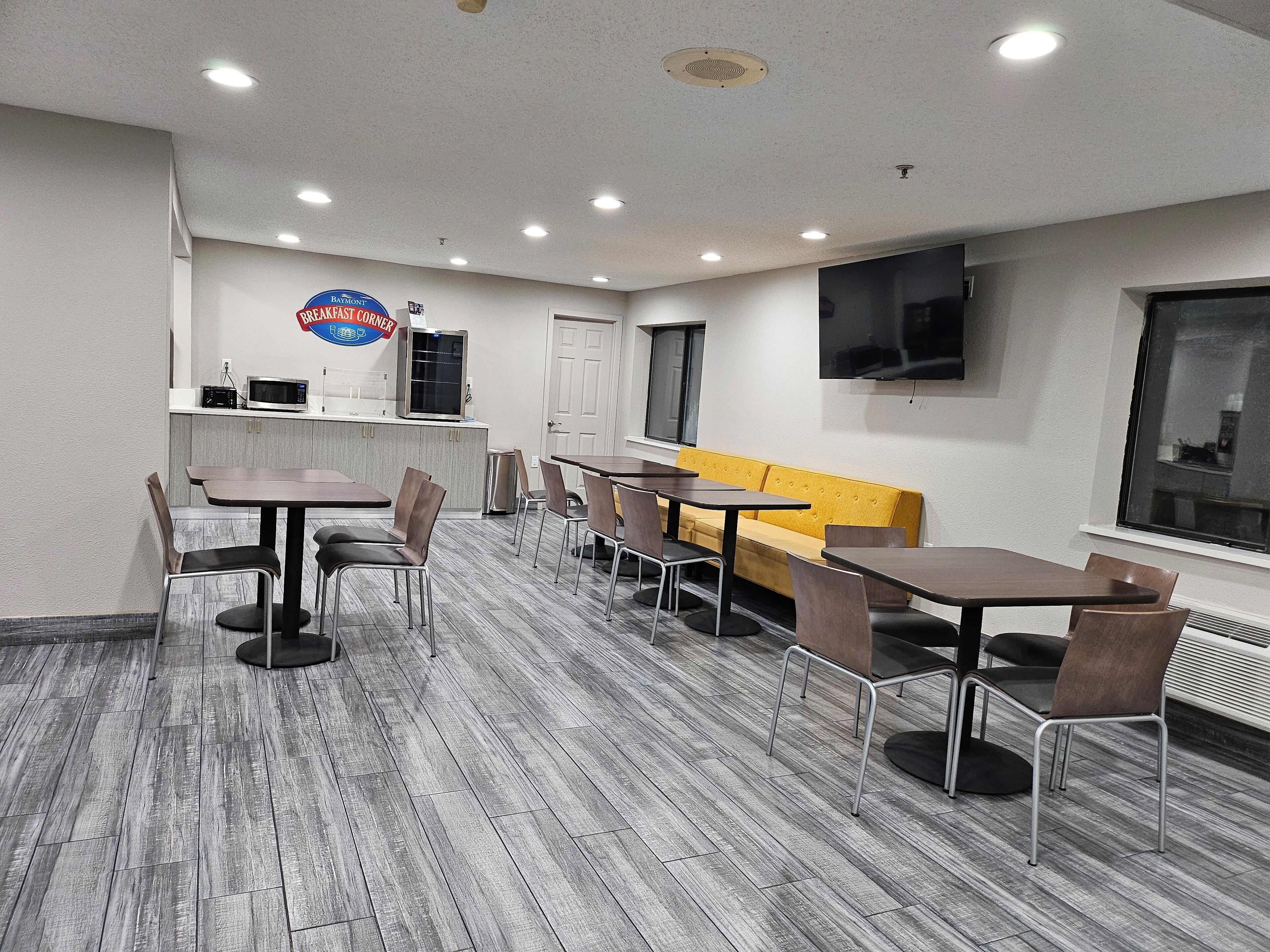 Un comedor moderno con varias mesas y sillas, un letrero de Baymont Breakfast Corner, un microondas y un televisor montado en la pared.