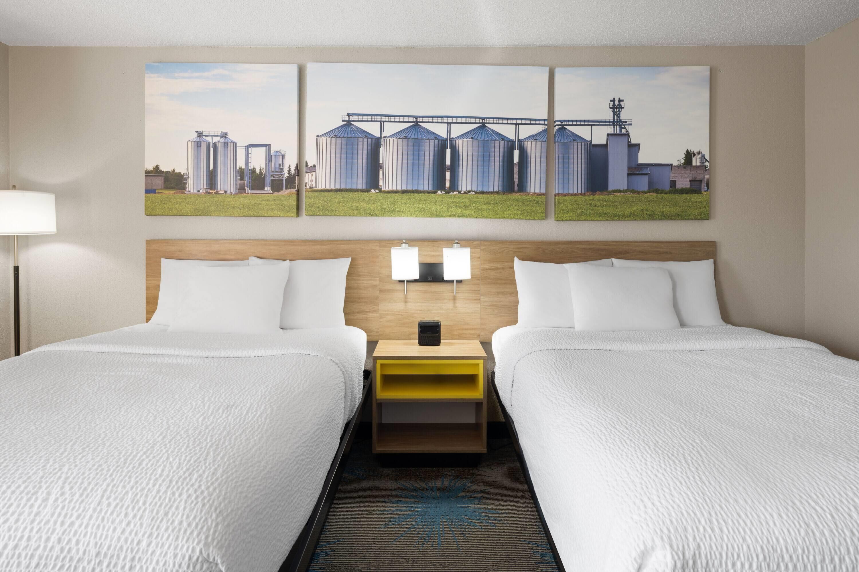 Habitación de hotel con dos camas, un cabecero de madera, una mesita de noche con lámparas y un cuadro de gran tamaño con temática industrial sobre las camas.
