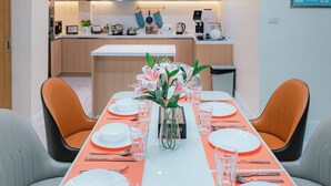 A modern dining area with a long table set for six, surrounded by orange chairs, a central vase with flowers, and a kitchen in the background.