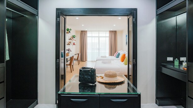 A hotel room with a glass partition, a bed with colorful pillows, a desk with a chair, and a wardrobe.