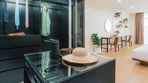 A modern hotel room with a glass desk, a hat on the table, a wardrobe with ties, a dining area with chairs and a table, and a bed.
