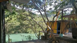 A covered outdoor area with a view of a river, a wooden deck, and a small structure with a blue door. - Room in Cabin - Rafting Hut by The River (San Agustin Lanquin)