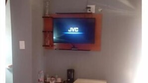 Un espacio habitable compacto con un televisor montado en la pared mostrando 'JVC'. Un refrigerador pequeño está apoyado contra la pared, y una cafetera sobre una repisa.
