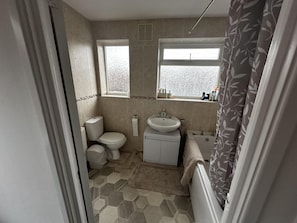 Baño con inodoro, lavamanos y bañera. Dos ventanas con vidrio esmerilado y cortina de ducha con estampado de hojas.